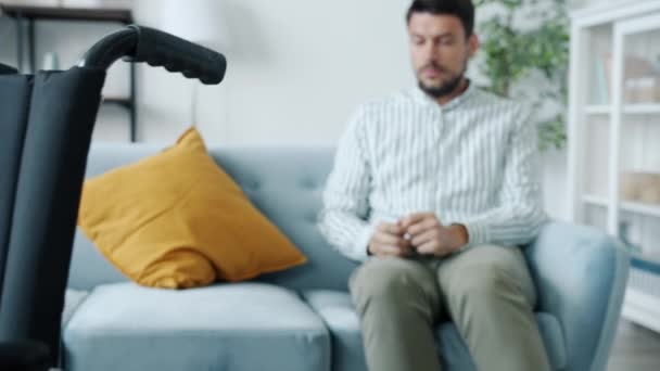 Zoom-in de homem com deficiência deprimido chorando sentado no sofá sozinho olhando para cadeira de rodas — Vídeo de Stock