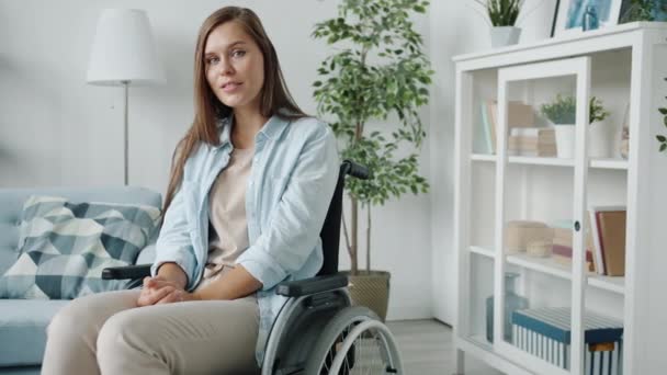 Portrét mladé ženy se zdravotním postižením sedí na invalidním vozíku doma a dívá se na fotoaparát s lehkým úsměvem — Stock video