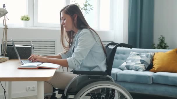 Motivated handicapped freelancer working with laptop typing and taking notes sitting in wheelchair at home — Stock Video