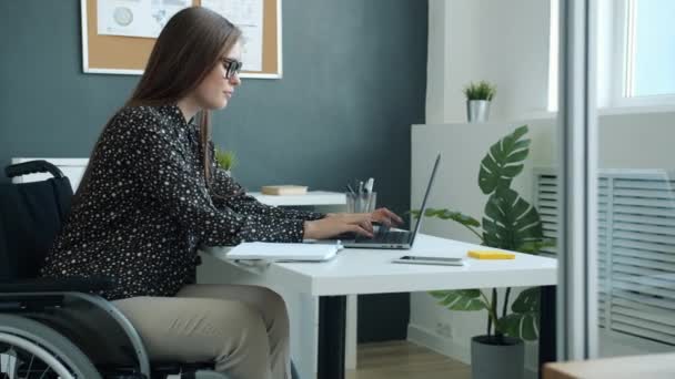 Jovem senhora de negócios com deficiência usando laptop digitação e escrita em notebook no escritório moderno — Vídeo de Stock