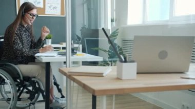 Özürlü ofis çalışanı bilgisayarla video görüşmesi yapıyor ve kulaklık takıp tekerlekli sandalyede oturuyor.