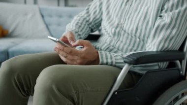 Tekerlekli sandalyedeki engelli adam yavaş çekimde akıllı telefon kullanarak evdeki ekranlara dokunuyor.
