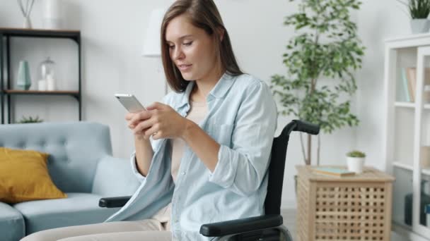 Krásná zdravotně postižená žena pomocí chytrého telefonu textování a usměvavé sezení na vozíku v bytě — Stock video