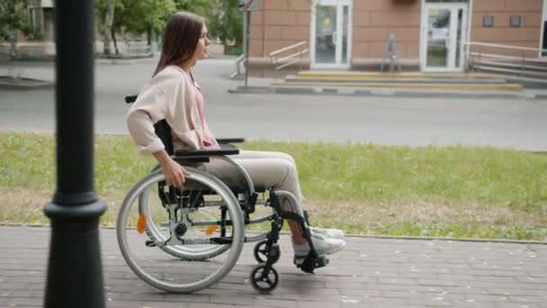 Vista lateral de una hermosa mujer discapacitada que se mueve en silla de ruedas a lo largo de la calle — Vídeos de Stock