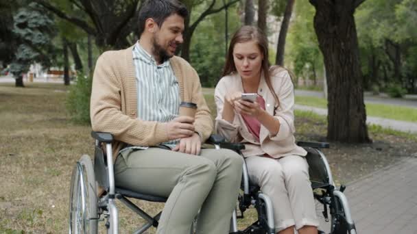 Pasangan cacat di kursi roda menggunakan ponsel pintar minum untuk minum kopi dan berbicara di luar ruangan — Stok Video