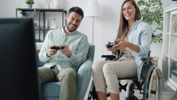 Joven hombre y mujer discapacitada en silla de ruedas jugando videojuegos hablando y riendo en casa — Vídeos de Stock