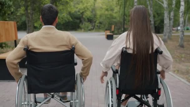 Back view of disabled people couple moving in wheelchairs in autumn park smiling enjoying fresh air — Stock Video