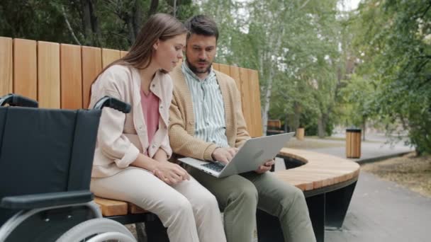 Movimiento lento de la pareja discapacitada usando el ordenador portátil y hablando en el banco al aire libre en el parque — Vídeo de stock