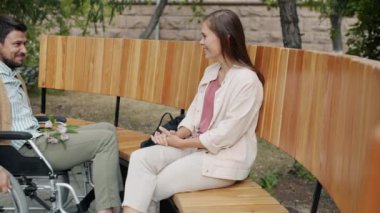 Tekerlekli sandalyedeki neşeli adam kız arkadaşıyla parkta buluşuyor. Çiçek getiriyor, konuşuyor, eğleniyor.