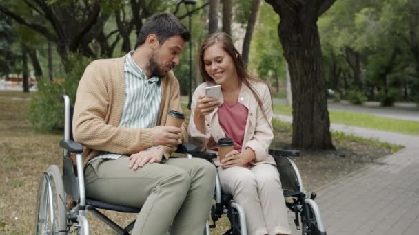 Deficiente marido e mulher sentado em cadeiras de rodas falando usando o smartphone beber café take-out no parque — Vídeo de Stock