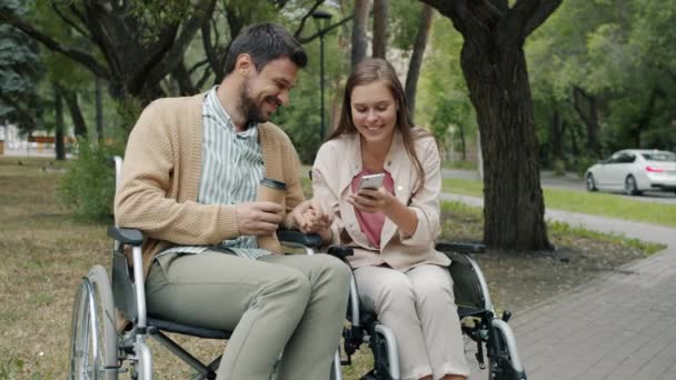 Movimento lento de jovens deficientes falando e usando smartphone sentado em cadeiras de rodas no parque — Vídeo de Stock