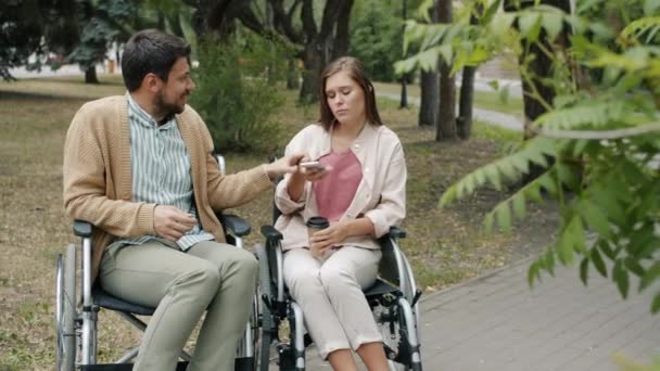 Funktionshindrade killen pratar med handikappade flicka utomhus tar sin smartphone sitter i rullstolar njuter park — Stockvideo