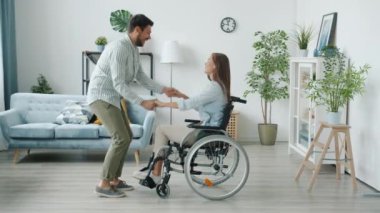 Tekerlekli sandalyede sakat genç bir kadın ve evde dans eden neşeli bir koca.