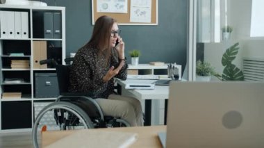 Engelli ofis çalışanı, tekerlekli sandalyede oturmuş cep telefonuyla konuşuyor. Tatsız sohbetlere odaklanmış.