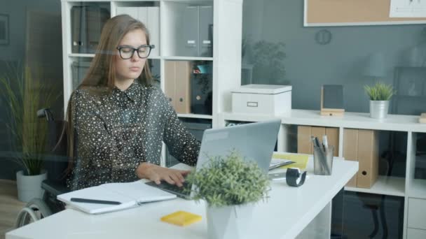 Empresaria cansada en silla de ruedas trabajando con portátil en la oficina y luego quitándose las gafas y tocando la cabeza — Vídeos de Stock