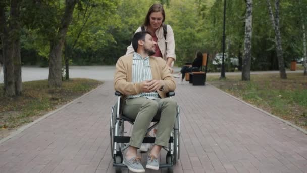 Jeune femme poussant fauteuil roulant avec mari handicapé parler souriant profiter de la promenade dans le parc — Video