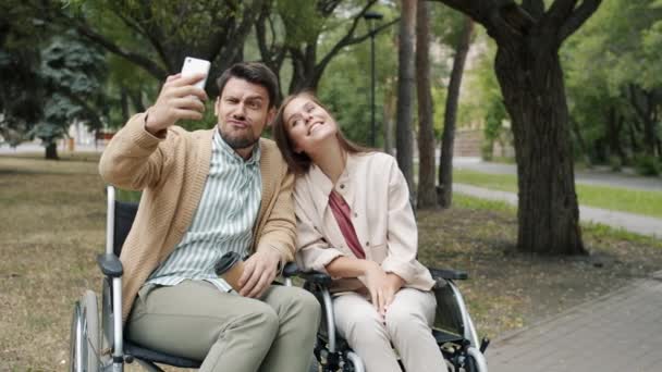 Fröhliches behindertes Paar macht Selfie mit Smartphone-Kamera und posiert mit lustigen Gesichtern im Rollstuhl im Park — Stockvideo