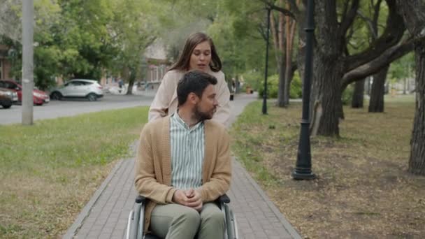 Movimento lento de cuidar jovem senhora empurrando homens cadeira de rodas falando sorrindo ao ar livre no parque — Vídeo de Stock