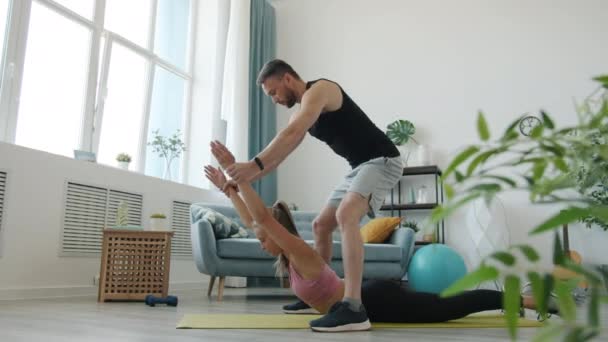 Behulpzaam vriendje ondersteuning van jonge dame tijdens stretching oefening doen sport binnen thuis — Stockvideo