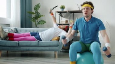 Dambıllarla egzersiz yapan komik bir adam video kaydeden ve elma yiyen bir arkadaş blogcuya spor topu fırlatıyor.
