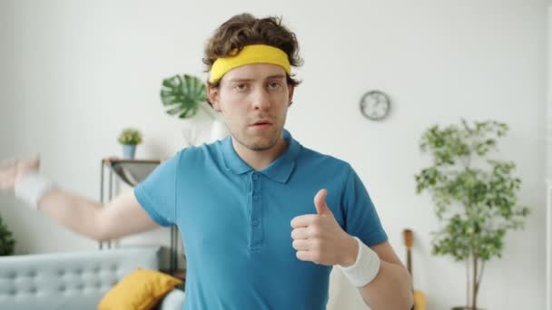 Porträtt av rolig ung man i sportkläder visar tummen upp hand gest och blinkande stående hemma — Stockvideo
