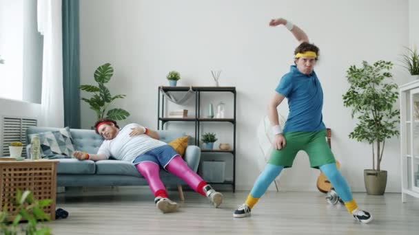 Zpomalený pohyb legrační mladý muž tančí a dělá bojová umění, zatímco přítel leží na pohovce dotýká srdce — Stock video
