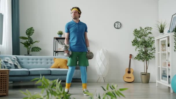 Lassú mozgás a gyenge srác színes vicces sportruházat emelés súlyzó és esik a kanapén otthon — Stock videók