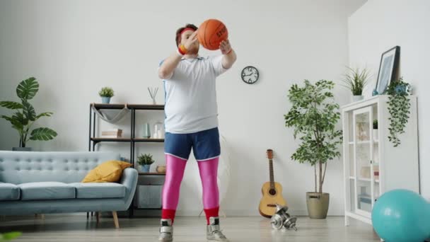 Zabawny młody człowiek w kolorowym stroju sportowym bawiący się koszykówką, a następnie wycelowany w kamerę i uśmiechnięty — Wideo stockowe