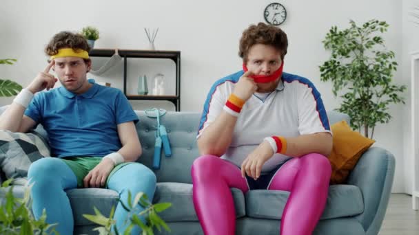 Нервові молоді чоловіки в ретро спортивному одязі, сидячи на дивані, відчувають тривогу і ненадійність в приміщенні вдома — стокове відео