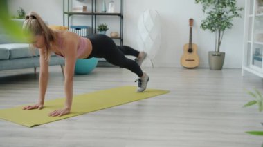 Formda genç bir kadın evde bireysel antrenman sırasında kollarını ve bacaklarını kaldırıyor.