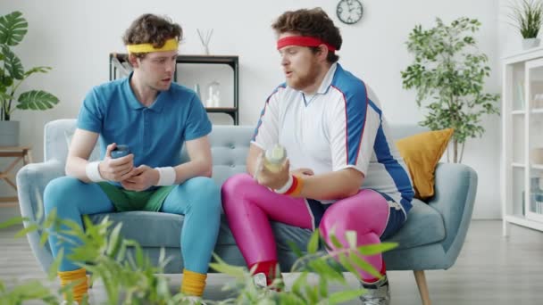 Божевільні хлопці, бодібілдери, займаються з гантелі і пляшкою з водою, розмовляючи про спорт, сидячи на дивані вдома — стокове відео