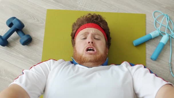 Вид сверху смешного лишнего веса парня, который упражняется, а потом ест хлеб, жуя багет, лежа на фитнес-коврике — стоковое видео