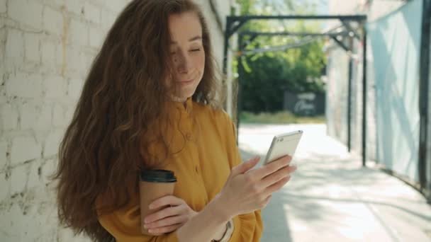 Mujer joven y guapa usando un teléfono inteligente sonriendo sosteniendo para ir a tomar un café afuera en la ciudad — Vídeo de stock