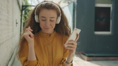 Kulaklık takan, açık havada müzik dinleyen, açık havada akıllı telefon ekranına bakan mutlu bir kız.