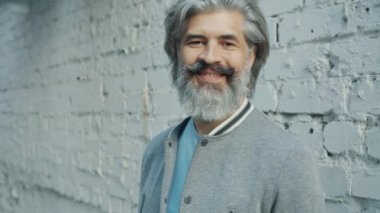 Arka planda tuğla duvarla ayakta gülümseyen çekici olgun bir adamın yavaş çekim portresi.
