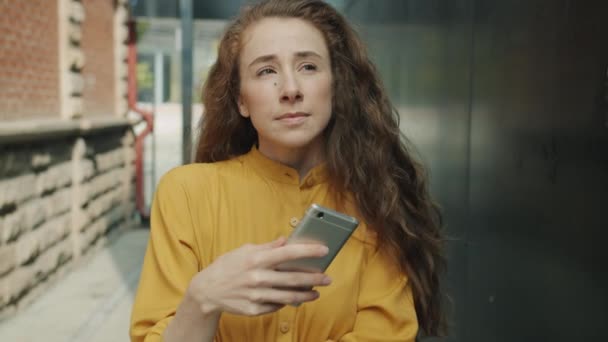 Movimento lento de menina atraente usando smartphone e sorrindo ao ar livre na rua da cidade — Vídeo de Stock