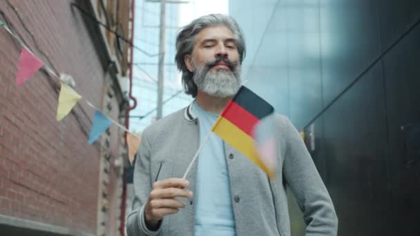 Retrato del atractivo hombre de pelo gris ondeando la bandera alemana y sonriendo afuera en la calle de la ciudad — Vídeos de Stock