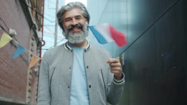 Movimiento lento de sonriente hombre maduro ondeando bandera nacional francesa de pie fuera de la ciudad moderna — Vídeos de Stock