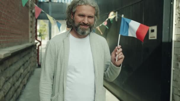 Медленный портрет счастливого француза, размахивающего национальным флагом, смотрящего в камеру и улыбающегося на городской улице — стоковое видео