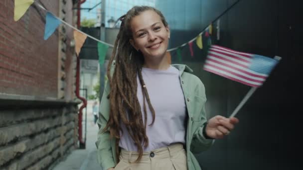 Ritratto di studentessa carina con dreadlocks in piedi fuori sventolando bandiera degli Stati Uniti e sorridendo guardando la fotocamera — Video Stock