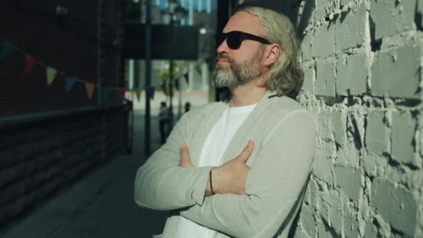 Retrato de homem elegante usando óculos de sol em pé ao ar livre com braços cruzados com parede de tijolo no fundo — Vídeo de Stock