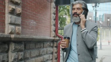 Yakışıklı, gri saçlı, cep telefonuyla konuşan ve dışarıda kahve içmek için içen adamın ağır çekimde görüntüsü.