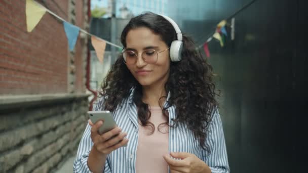 Slow motion van gelukkig aziatisch meisje in hoofdtelefoon genieten van muziek met behulp van smartphone en dansen buiten — Stockvideo