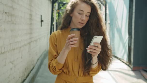 Lambat gerak wanita muda riang berjalan menggunakan telepon pintar dan minum untuk pergi kopi di luar ruangan di kota — Stok Video