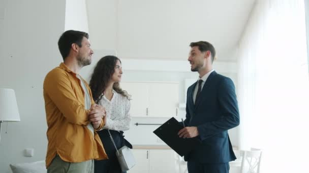 Щасливі клієнти чоловік і дружина купують будинок, отримуючи ключ від агента в приміщенні в світлій кімнаті — стокове відео