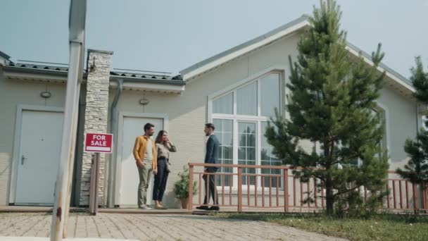 Joyful couple talking to real estate agent standing near beautiful suburban house outdoors — Stock Video