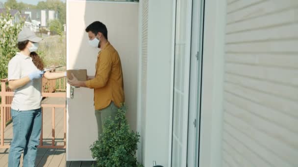 Man in medical mask receiving package from delivery woman signing paper during covid-19 lockdown — Stock Video