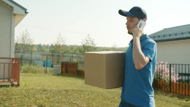 Tipo de entrega en cámara lenta hablando por teléfono móvil llevando parcela en el área suburbana — Vídeos de Stock