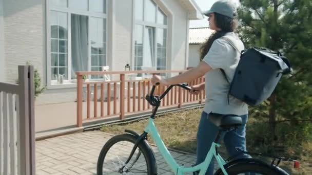 Levering werknemer meisje met rugzak paardrijden fiets dan kloppen op klanten deur — Stockvideo