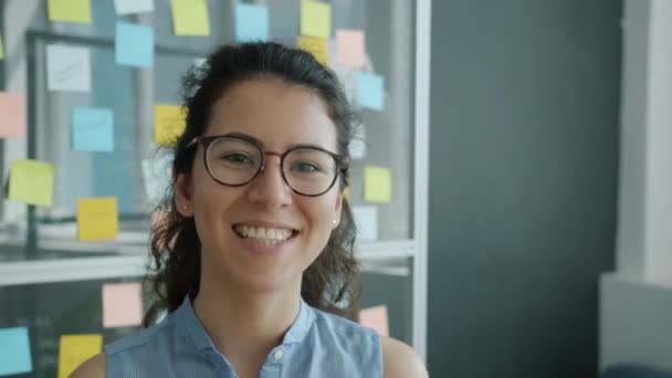 Retrato en cámara lenta de la atractiva joven morena en gafas de pie en el interior de la oficina creativa sonriendo — Vídeos de Stock